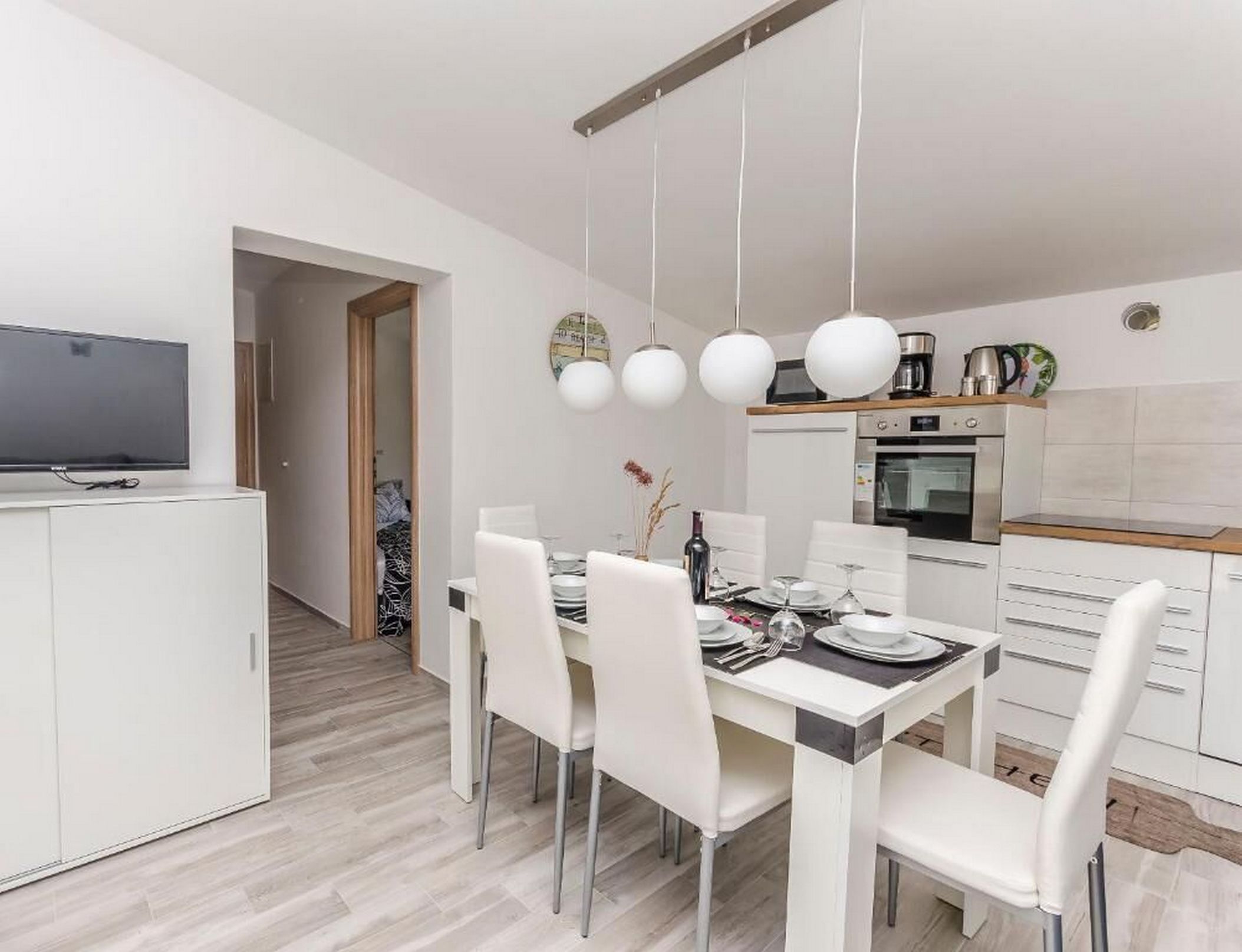 Una cocina moderna con muebles blancos, una mesa de comedor lista para cuatro personas y un televisor de pantalla plana montado en la pared.