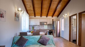 A bedroom with a bed, a wooden ceiling, a kitchen with cabinets and appliances, and a bathroom visible through an open door. - House Olive Tree (Rab)