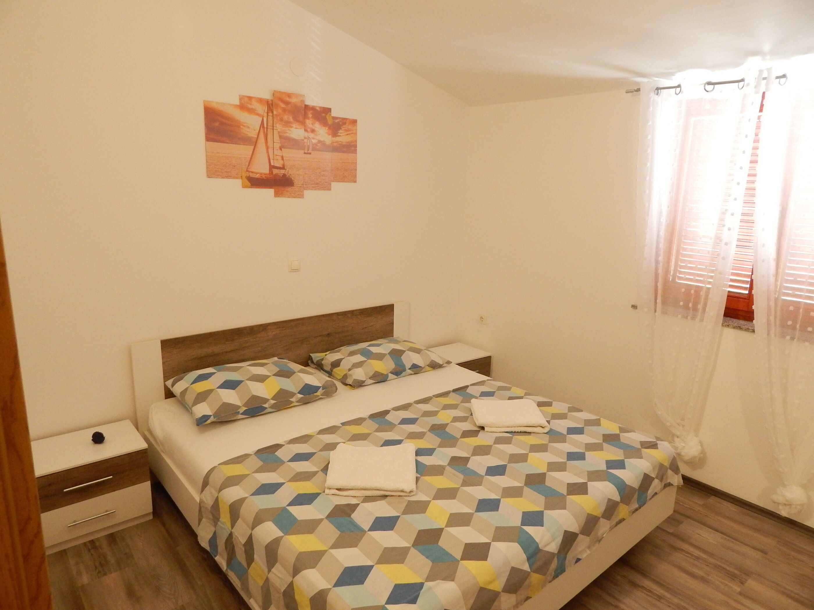 A bedroom with a bed, a nightstand, a drawer unit, a wall-mounted picture, and a window with curtains.