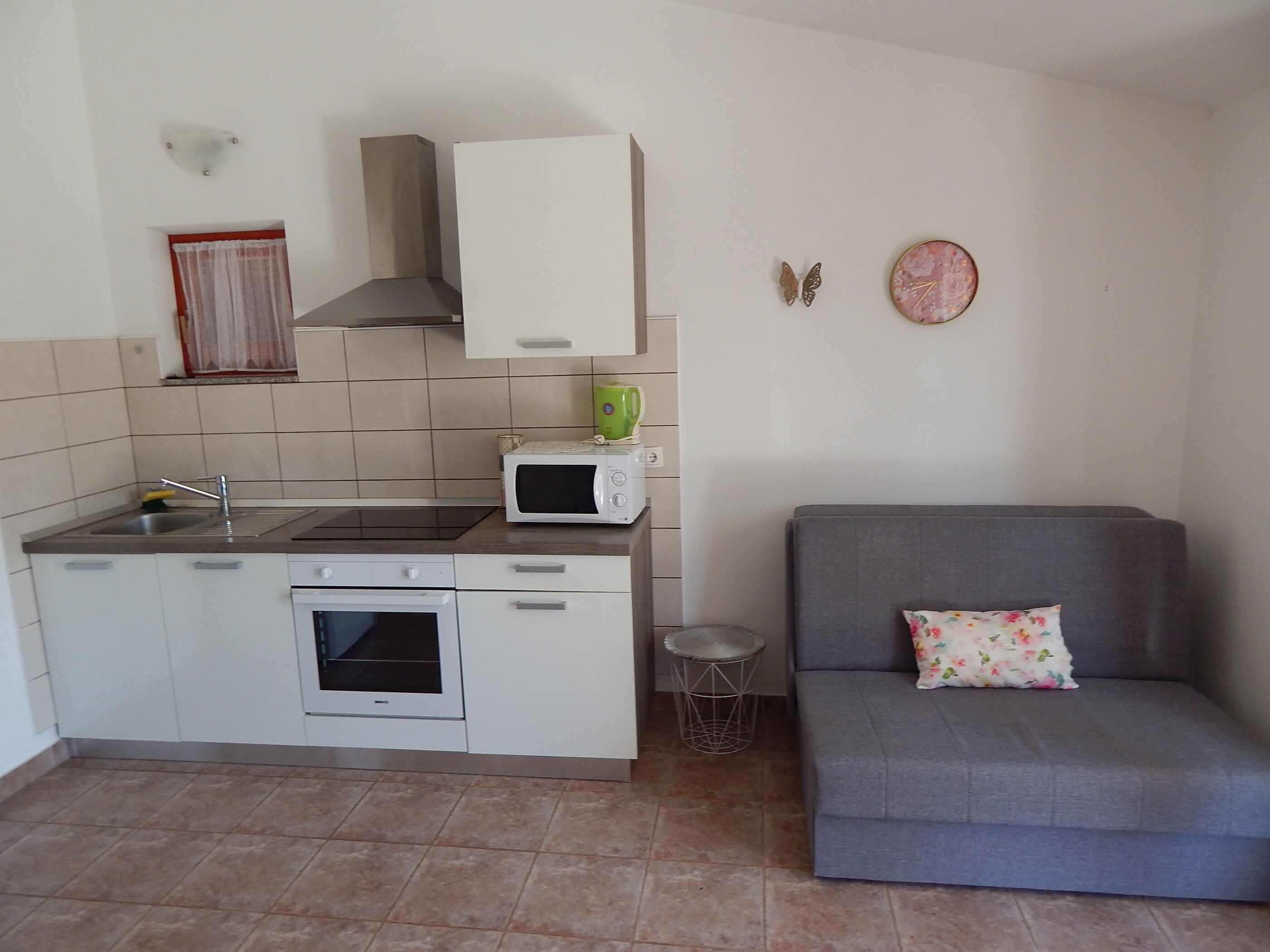 A compact kitchen with a microwave, stove, and sink. A grey sofa with a floral pillow is against the wall.