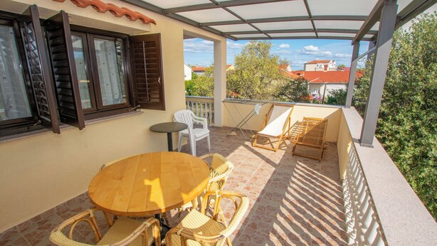 A covered outdoor patio with a round wooden table and wicker chairs, a small round table, and a folding chair. - Apartments Barbara (Pakostane)