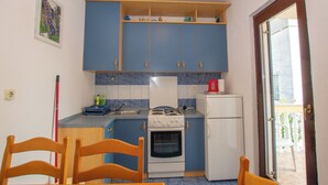 A compact kitchen with blue cabinets, a stove, a refrigerator, and a dining area with wooden chairs. - Apartments Barbara (Pakostane)