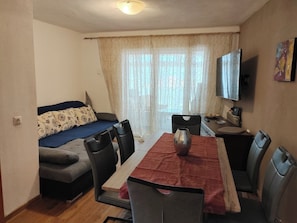 A compact living space with a sofa, dining table, chairs, a flat-screen TV, and a window with sheer curtains. - Apartment Kristina (Crikvenica)