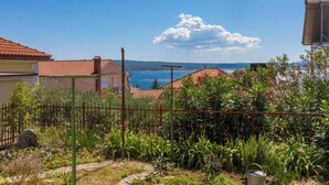 Ein Garten mit einem Metall-Gießkann, einem Kiesweg und im Hintergrund Blick auf das Meer und die Berge.