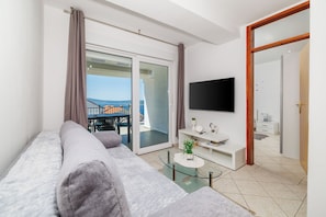 A modern living room with a sofa, a flat-screen TV, a glass coffee table, and a sliding glass door leading to an outdoor area. - Apartments Grozdana (Crikvenica)