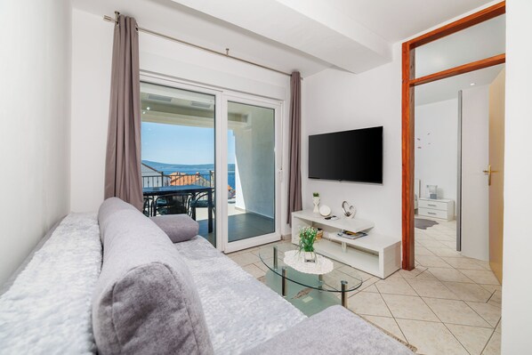 Una sala de estar moderna con un sofá, un televisor de pantalla plana, una mesa de centro de vidrio y un puerta corrediza de cristal que da a un espacio exterior.
