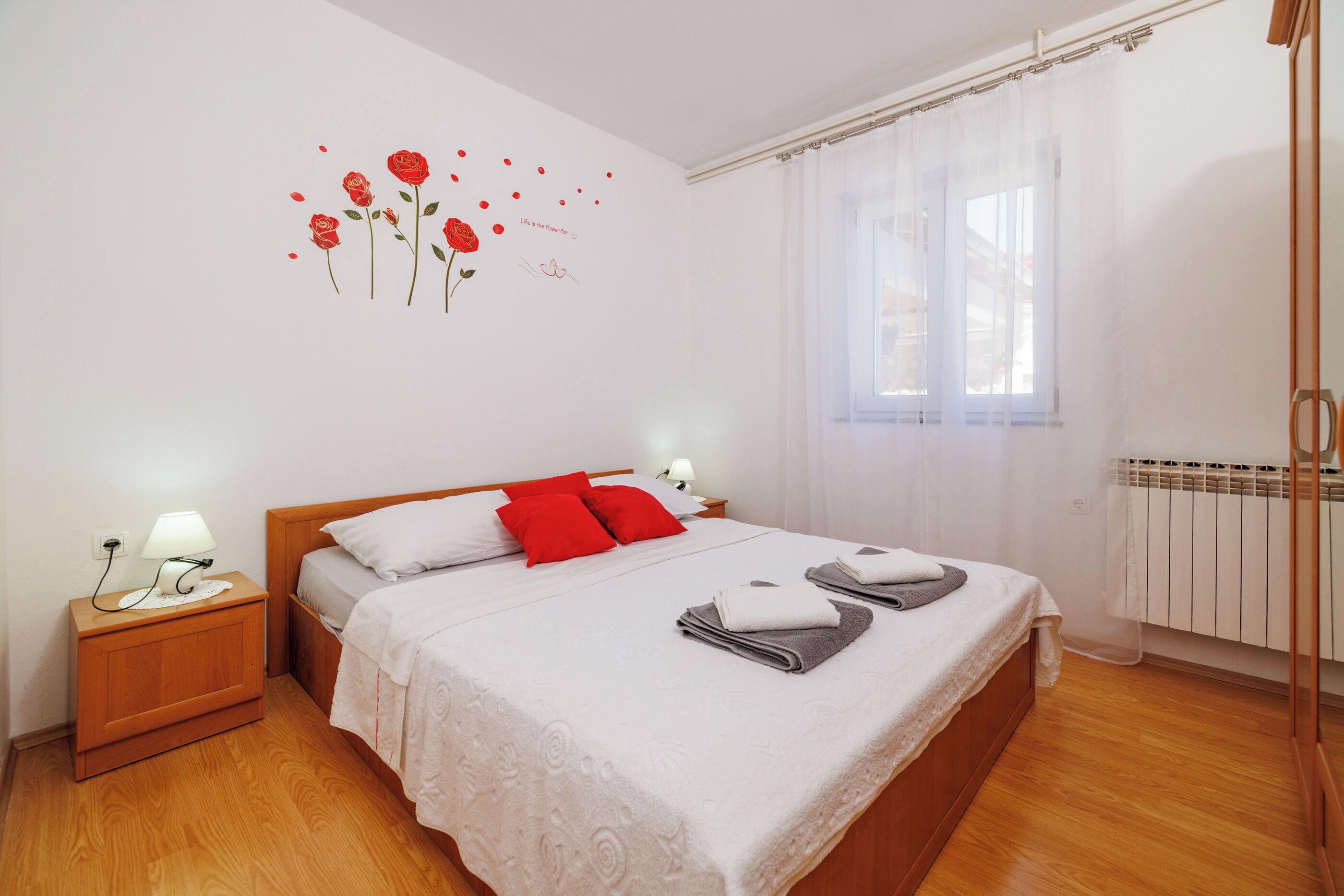 A neatly made bed with red pillows, a bedside table with a lamp, and a wall decoration of roses.