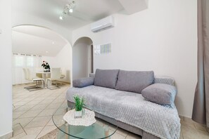A modern living room with a grey sofa, a glass coffee table with a plant, and a dining area in the background. - Apartments Grozdana (Crikvenica)