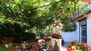 Un área de comedor al aire libre con una mesa larga dispuesta para cuatro personas, rodeada de plantas en macetas y una pérgola sobre ella.