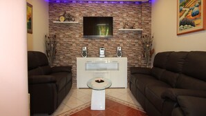 Una sala de estar con dos sofás oscuros, un mueble blanco para entretenimiento con altavoces, un televisor de pantalla plana y una pared con diseño de ladrillo.