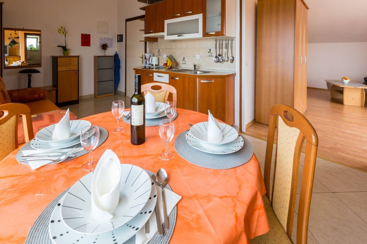 Een eetgedeelte met een tafel voor vier personen, een keuken op de achtergrond en een zithoek met een bank en een lamp.