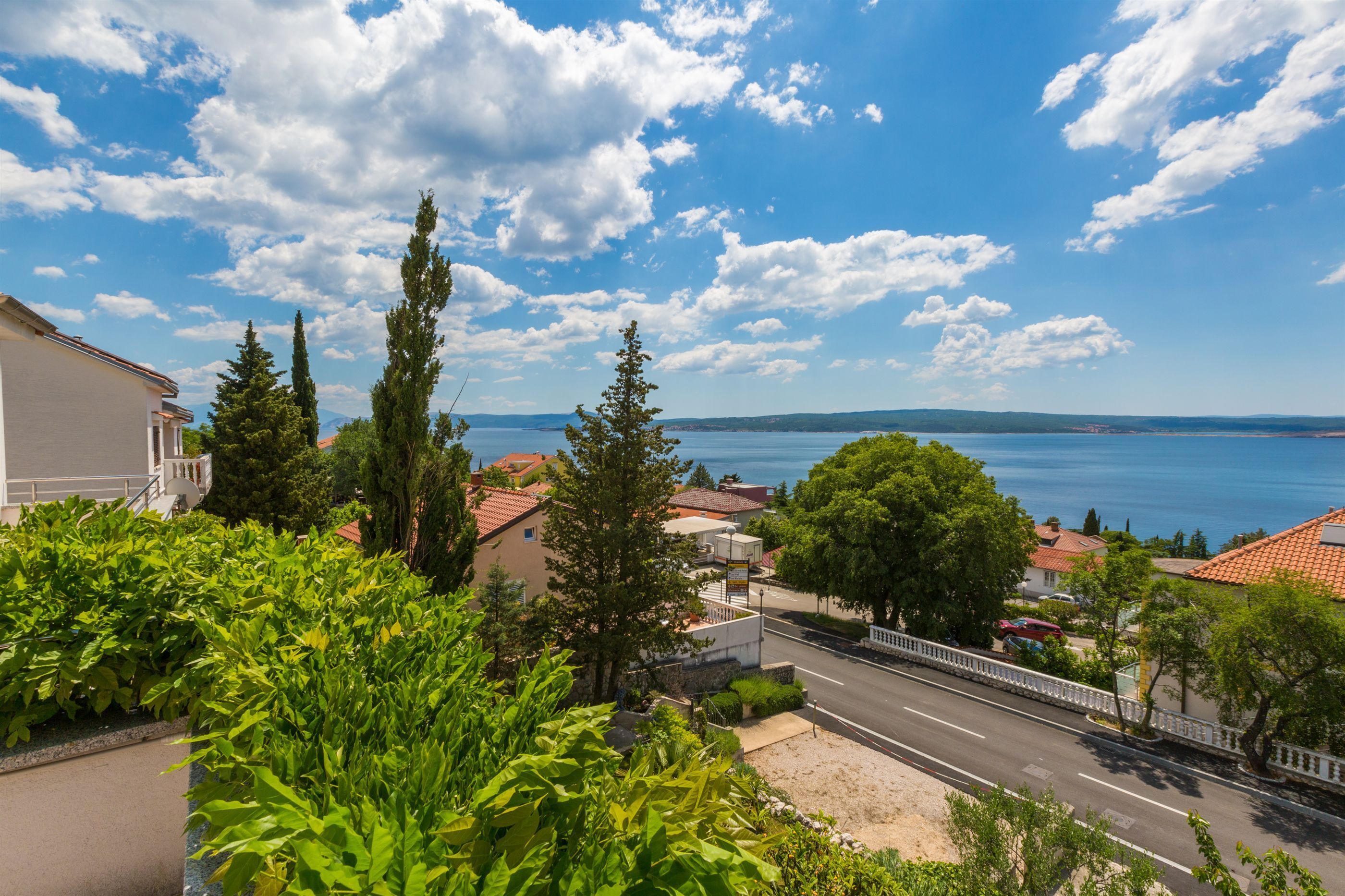 Une ville côtière offrant une vue dégagée sur la mer, une rue bordée de voitures garées et des immeubles résidentiels aux toits recouverts de tuiles rouges.