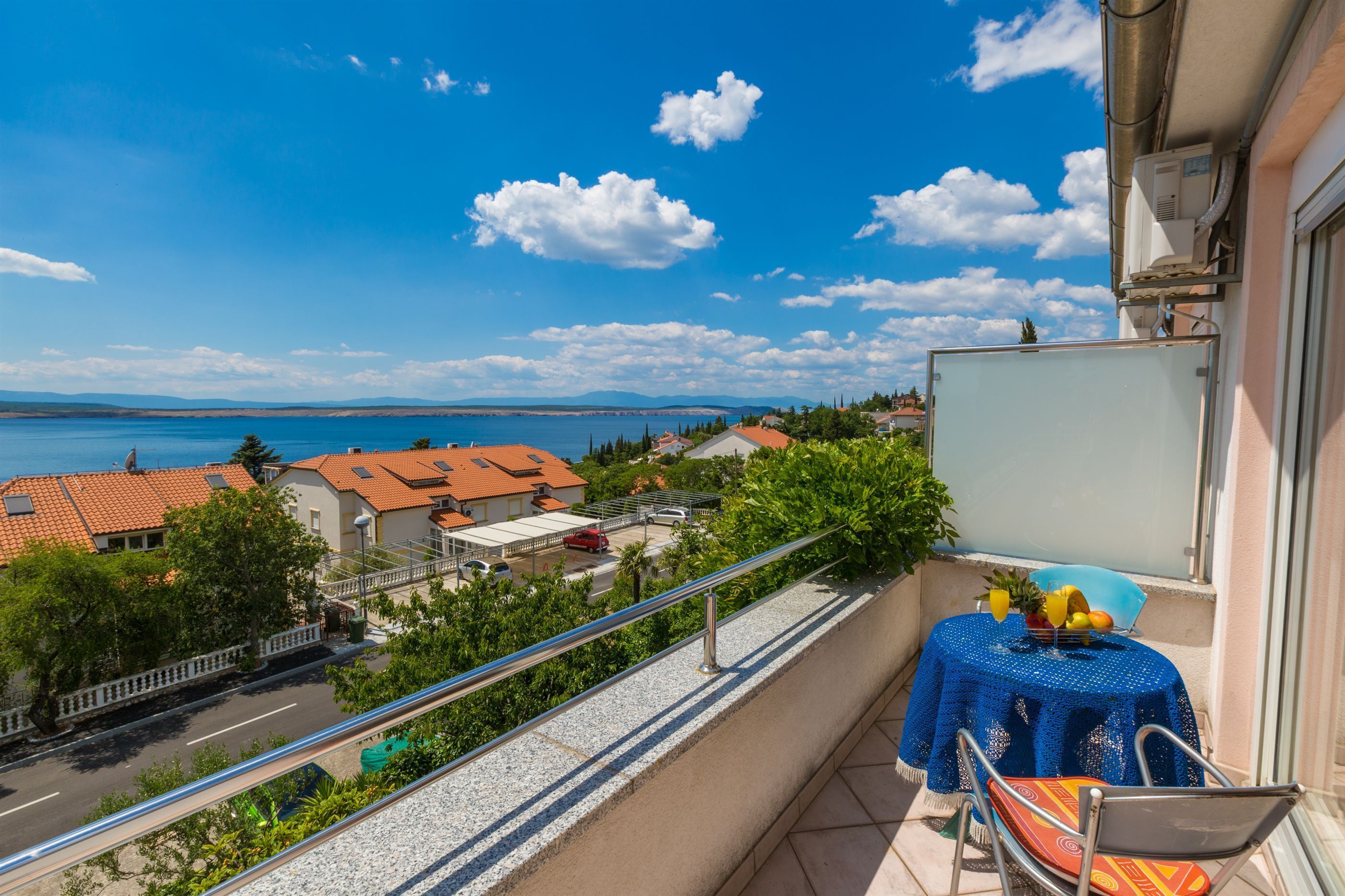 En balkon med et bord og en stol, udsigt over et kystområde med huse og en klar himmel.