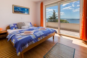A bedroom with a bed, a nightstand, a lamp, a window with curtains, and a view of the sea. - Apartments Vanessa (Crikvenica)
