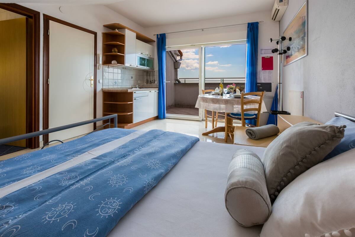 Een compacte hotelkamer met een bed, een kleine kitchenette, een zithoek met een tafel en stoelen, en een balkon met uitzicht.