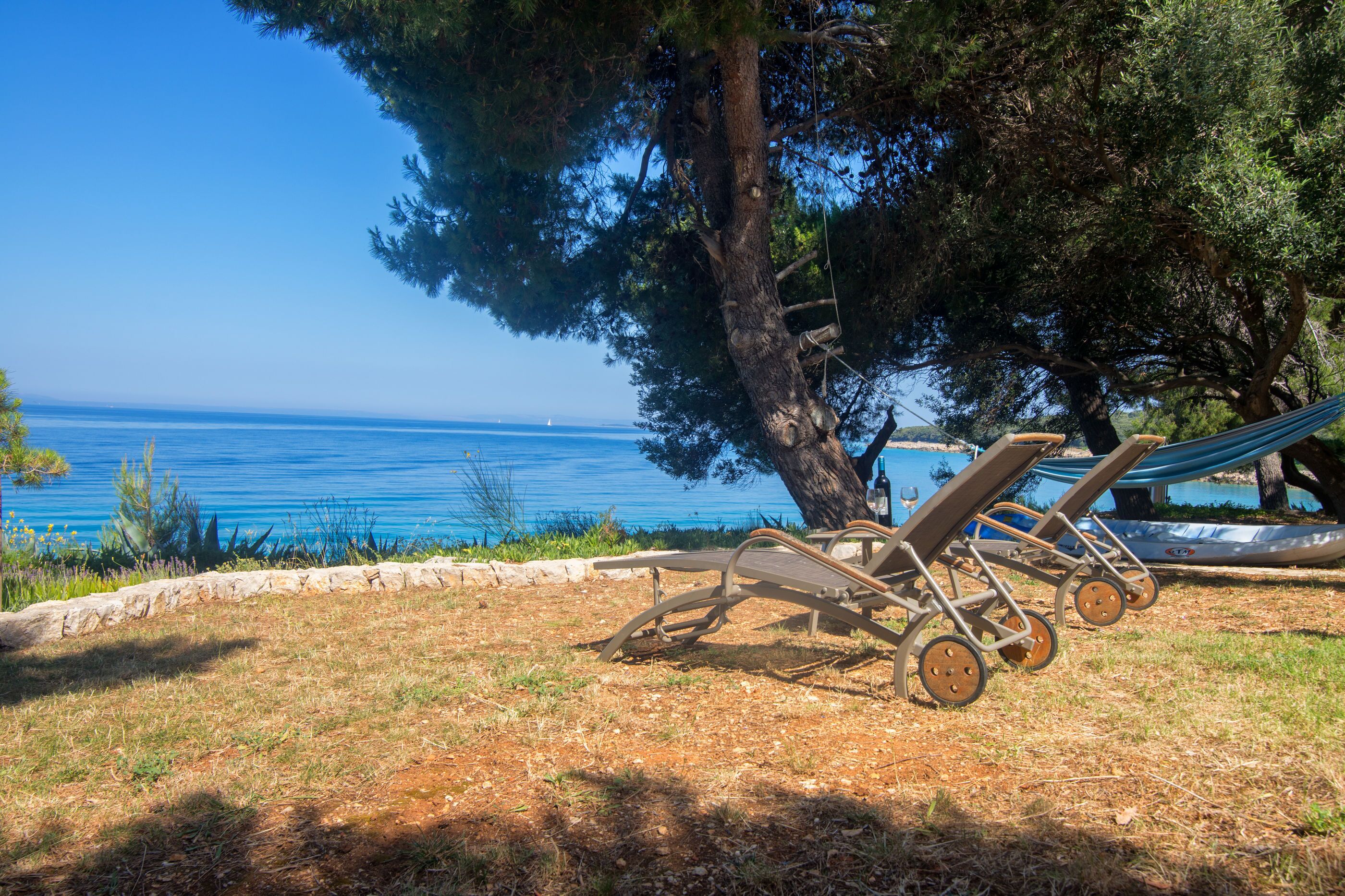 A beachside setting with a hammock and two sun loungers under a large tree, overlooking the sea.