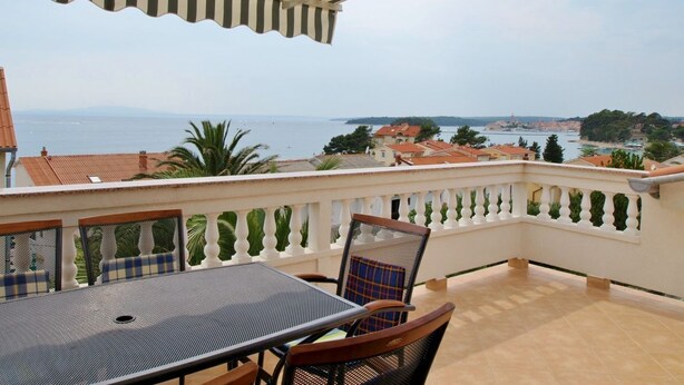 Eine Terrasse mit Tisch und Stühlen, ein gestreifter Sonnenschirm und Blick auf das Meer und Gebäude.