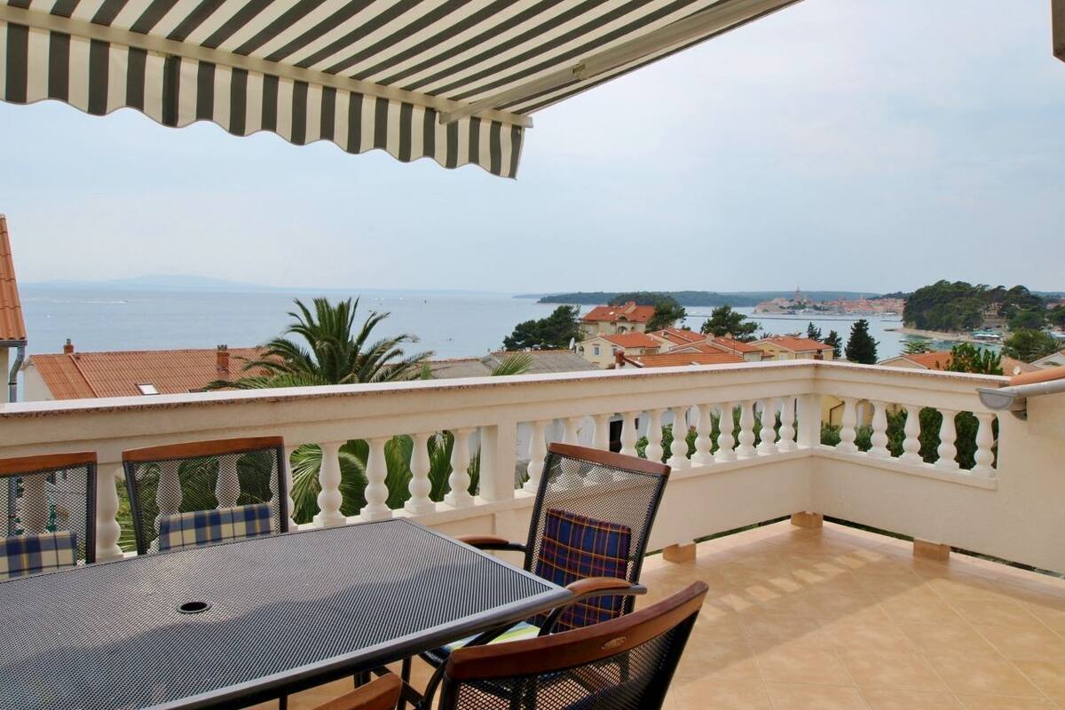 Eine Terrasse mit Tisch und Stühlen, ein gestreifter Sonnenschirm und Blick auf das Meer und Gebäude.