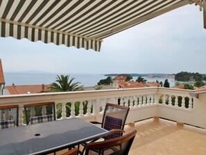 Eine Terrasse mit Tisch und Stühlen, ein gestreifter Sonnenschirm und Blick auf das Meer und Gebäude.