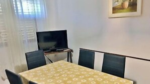 Ein Essbereich mit einem Tisch, der mit einer blumigen Tischdecke bedeckt ist, umgeben von Stühlen. Über dem Tisch ist ein Fernseher an der Wand befestigt.
