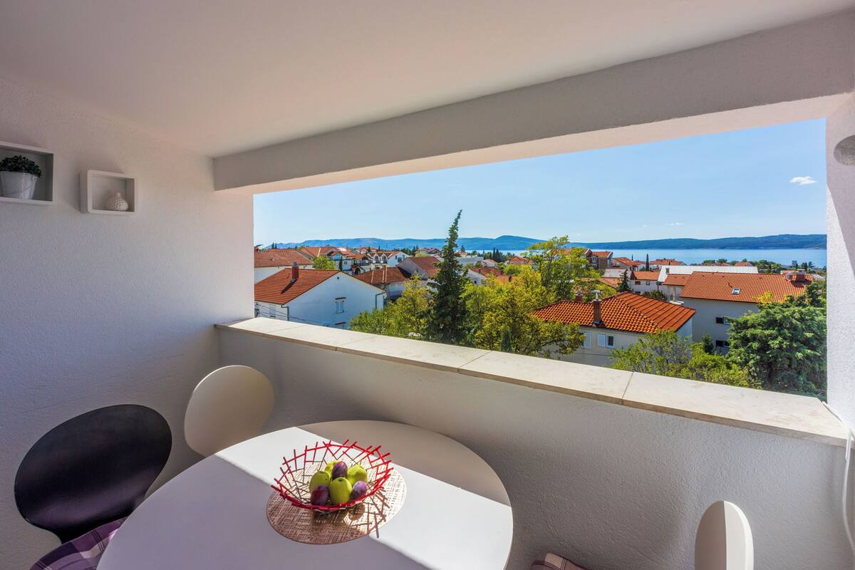 A balcony with a table and chairs, a bowl of fruit, and a view of a residential area with red-roofed houses and a body of water in the distance.