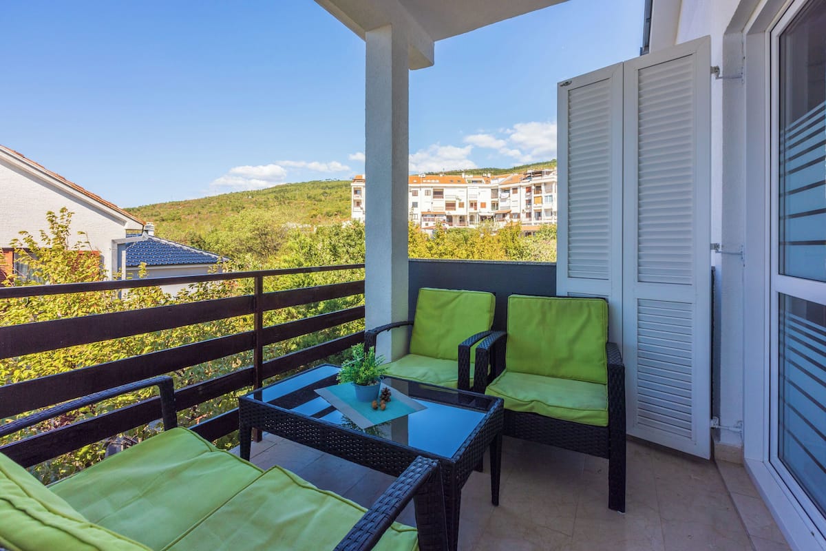 A balcony with wicker furniture, a glass table, and a view of buildings and greenery.