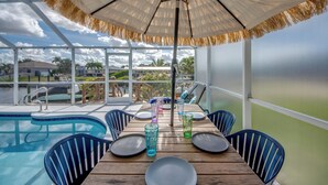 Et spiseområde ved poolen med et bord til fire personer, omgivet af stole, og en parasol der giver skygge.