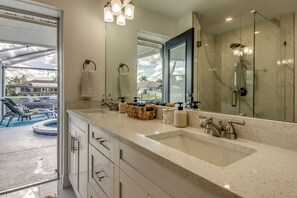 Un baño con un espejo grande, dos lavamanos y una ducha. Hay una ventana con vistas a una piscina y casas.