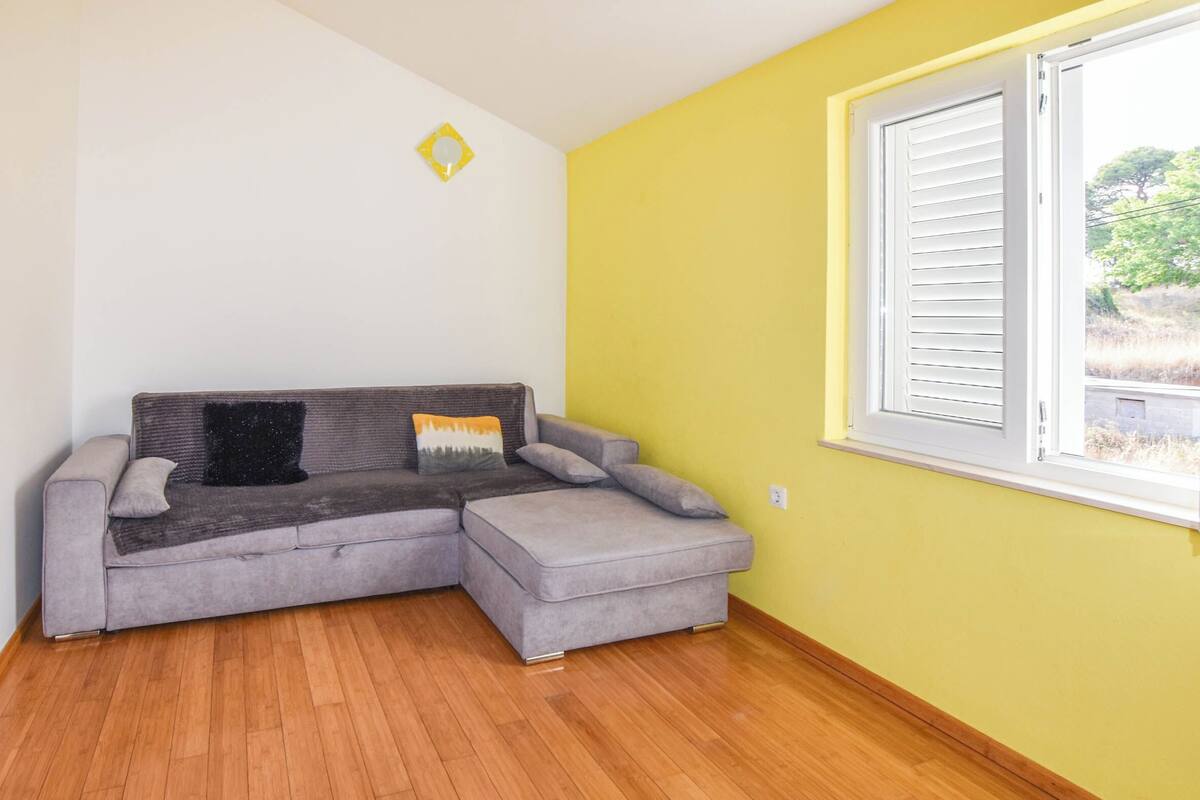 Une pièce avec un canapé d’angle gris, un mur jaune et une fenêtre avec des stores.