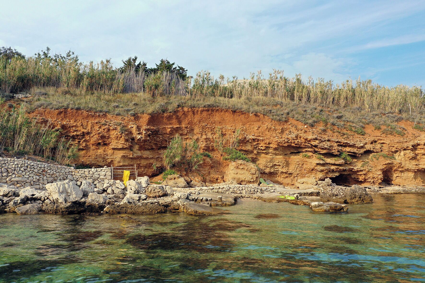A rocky shoreline with clear water, a stone wall, and sparse vegetation.