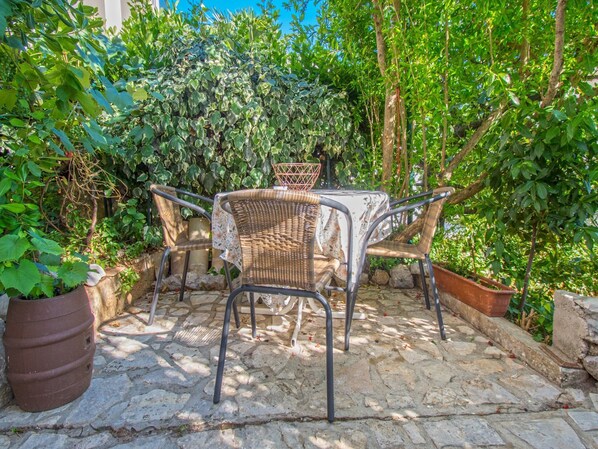 An outdoor patio with a table and four wicker chairs, surrounded by lush greenery and stone flooring. - Apartments Stella (Crikvenica)