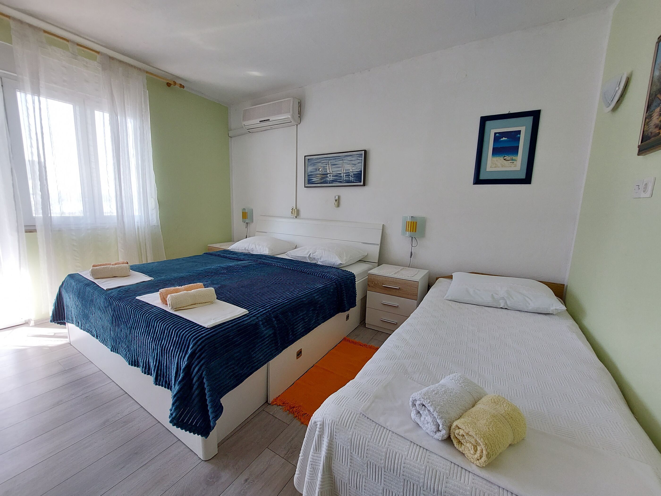 A hotel room with two beds, a nightstand, a wall-mounted air conditioner, and a window with sheer curtains.