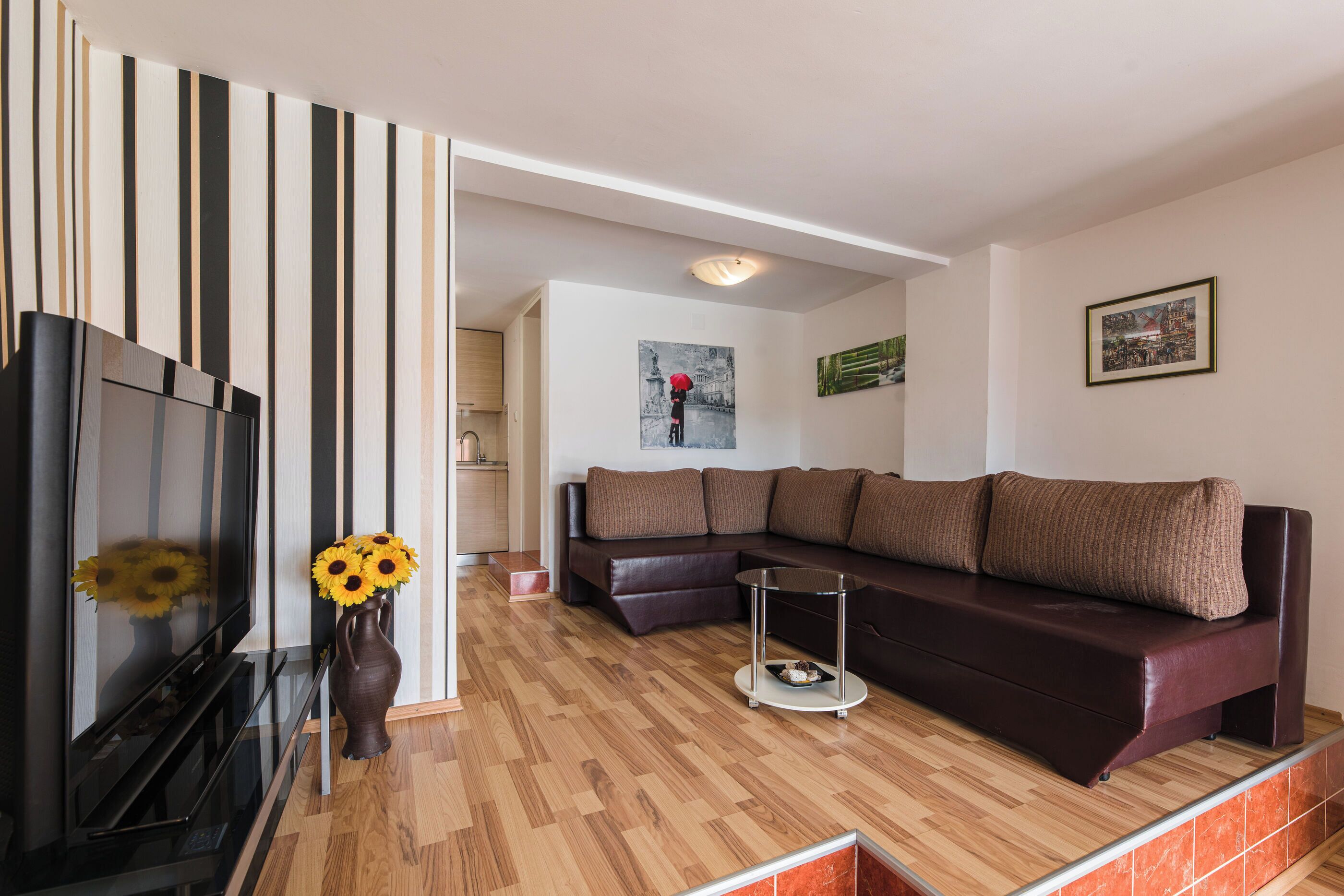 A modern living room with a sectional sofa, a flat-screen TV, a vase with sunflowers, and a wooden floor.