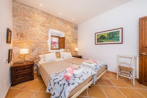 A bedroom with a stone wall, a bed with a patterned bedspread, a wooden chair, and a painting on the wall.