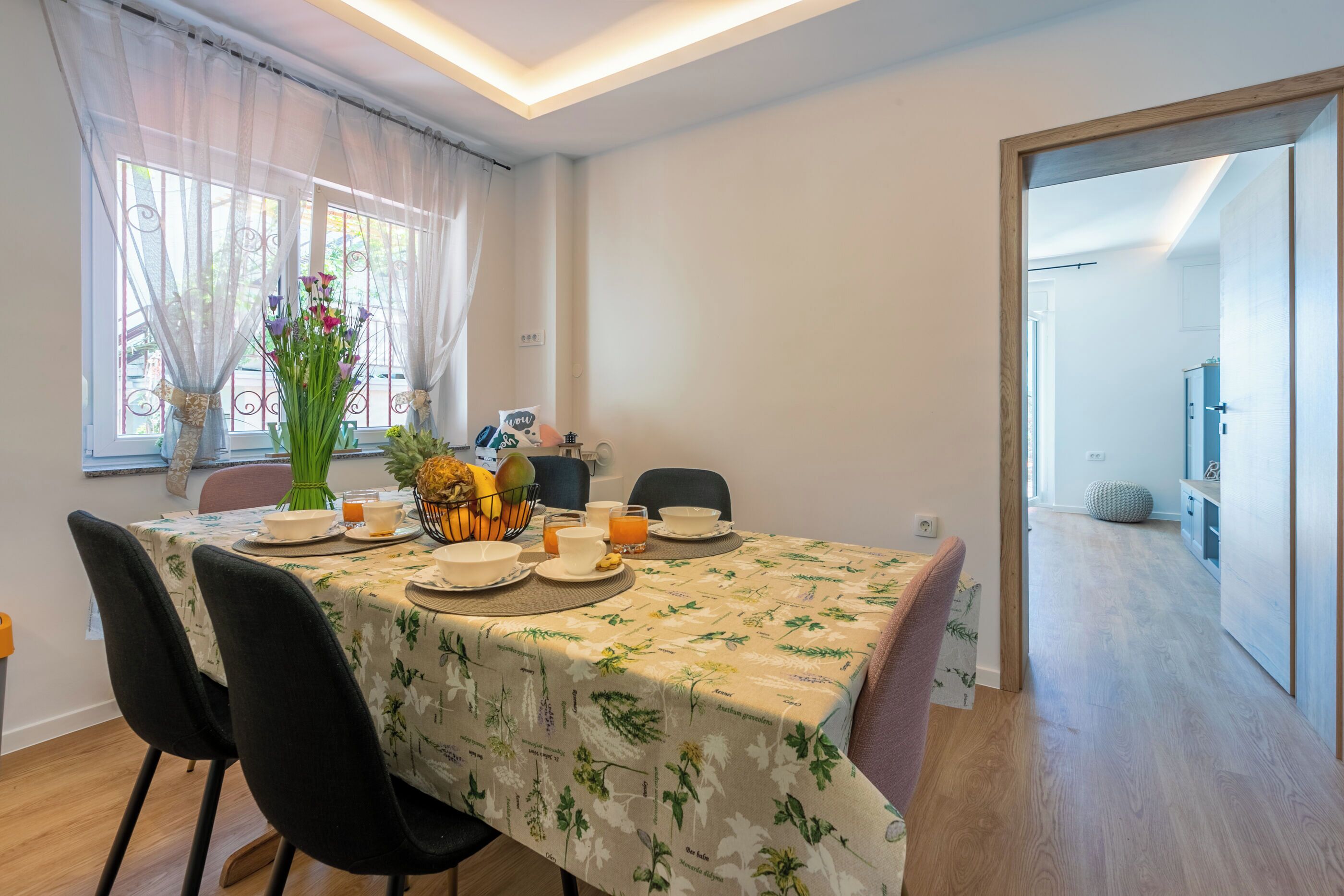 A dining area with a table set for four, a vase with flowers, and a fruit basket.