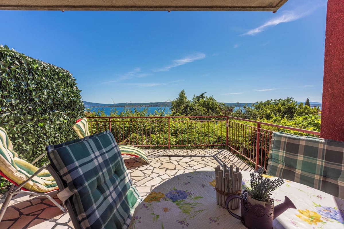 Un balcone con vista sul mare, un tavolo con tovaglia fiorata e due sedie con cuscini a quadri.