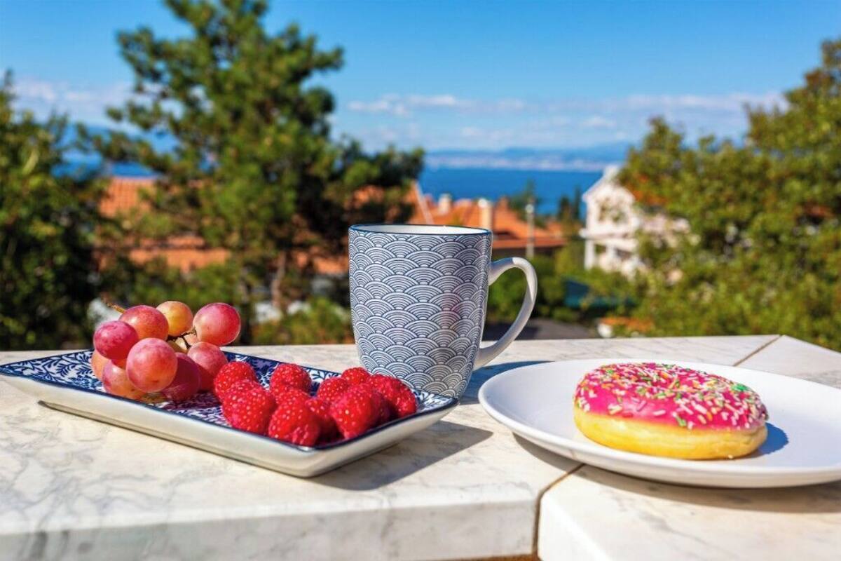 A table with a plate of strawberries and a plate of pink frosted doughnut, a patterned mug, and a scenic view in the background.