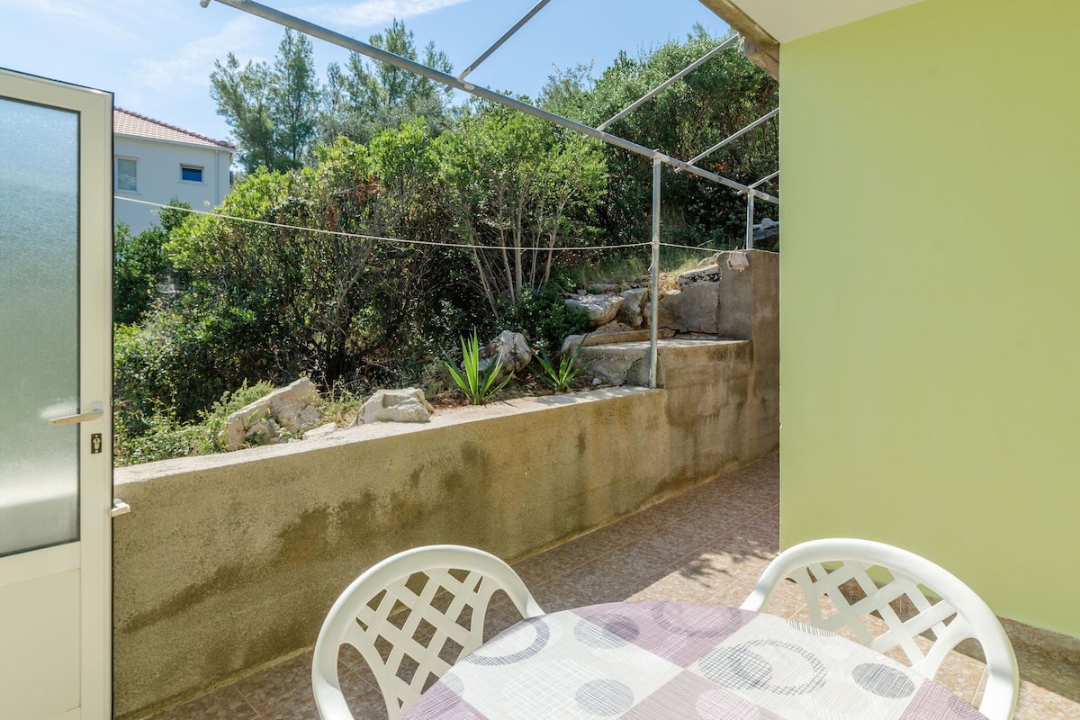 Un piccolo patio con tavolo e due sedie, un muro in cemento e vista su un giardino con alberi e rocce.