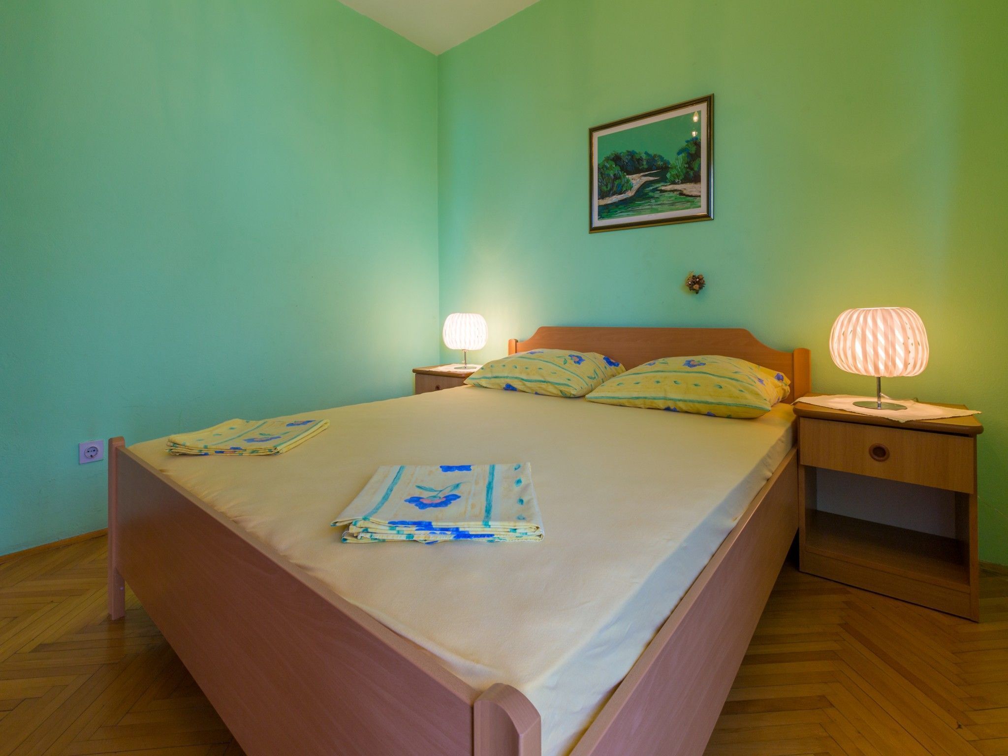 A bedroom with a bed, two bedside tables, a nightstand, a wall-mounted lamp, and a framed picture.