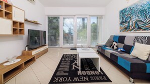 A modern living room with a flat-screen TV, a sofa, a coffee table, and a large window with blinds. - Apartment Villa Sara (Crikvenica)