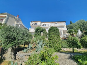 Un edificio residencial de varios pisos con balcones, rodeado de abundante vegetación y un muro de piedra.
