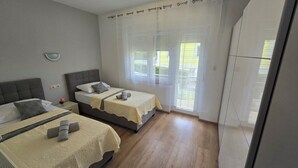Une chambre avec deux lits, une petite table de chevet avec une lampe, une étagère fixée au mur et une grande fenêtre avec des rideaux.