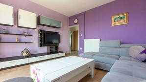 Un salon avec un canapé gris, une table basse blanche, un téléviseur à écran plat fixé au mur et un mur violet.