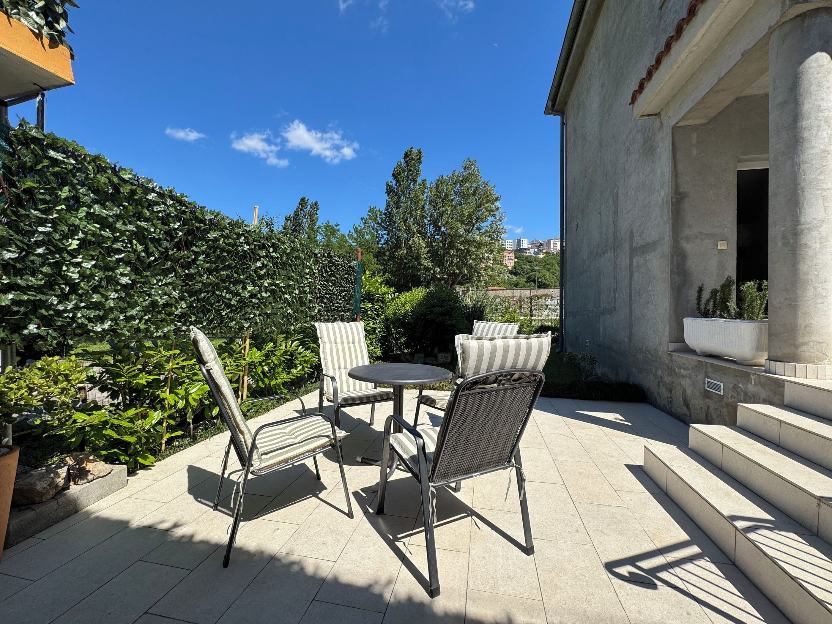 An outdoor patio with a table and chairs, surrounded by greenery and a view of buildings in the distance.