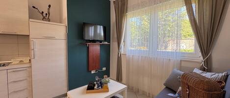 A compact living space with a kitchenette, a flat-screen TV mounted on a wooden shelf, a dining table, a sofa with a woven basket and a hat, and a window with sheer curtains.