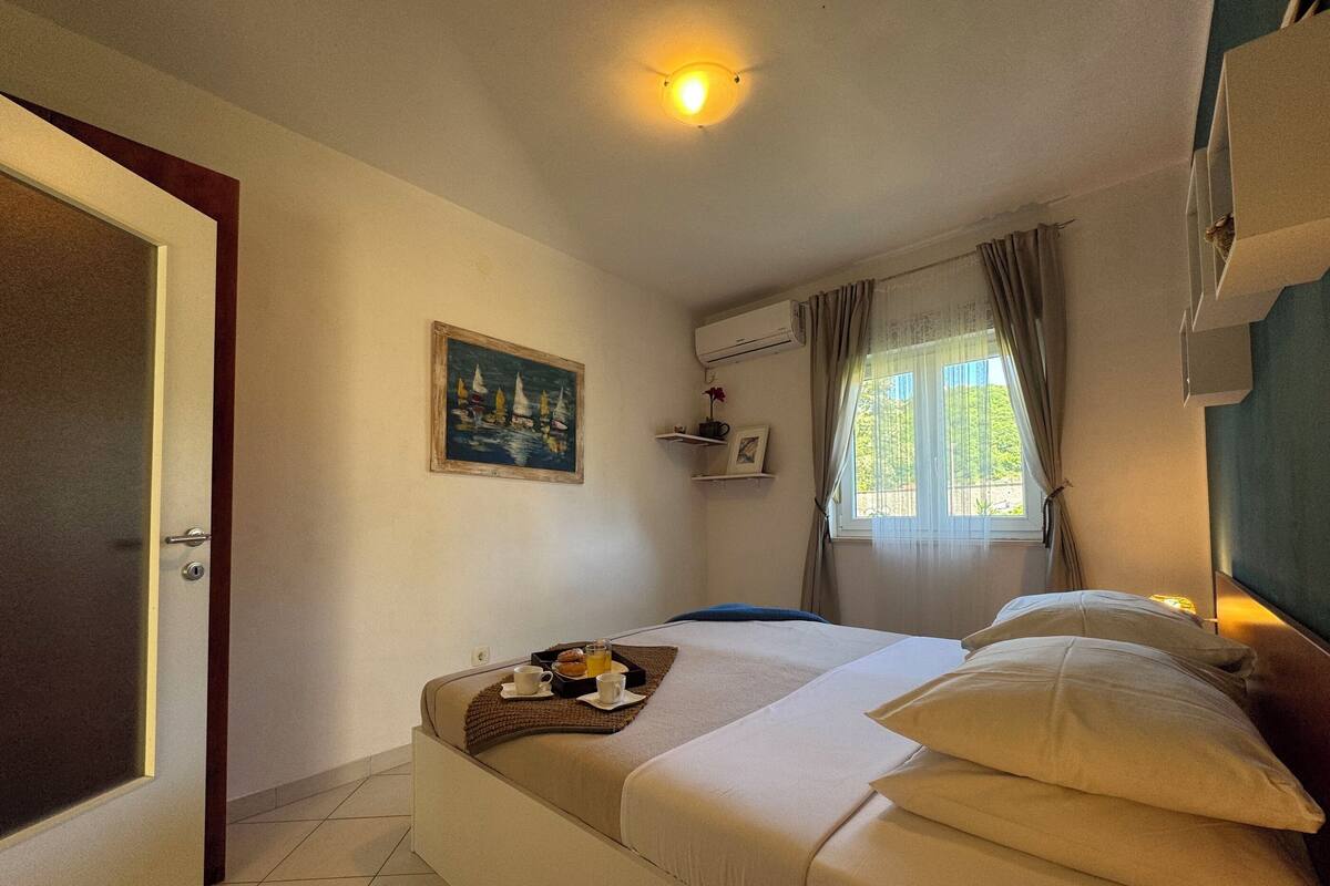 A bedroom with a bed, a painting, an air conditioning unit, a window with curtains, and a door.