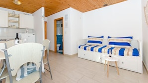 Un espacio habitable compacto con kitchenette, una zona de comedor con una silla, un sofá con ropa de cama a rayas y una mesita con una botella.