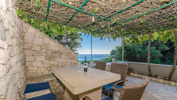 Un espacio al aire libre con paredes de piedra, una mesa y sillas, una pérgola arriba y vistas al mar.