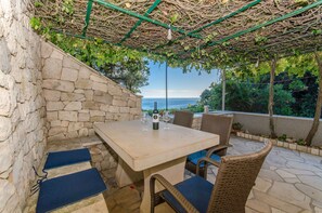 A stone-walled outdoor seating area with a table and chairs, a pergola overhead, and a view of the sea. - House ULE (Korčula)
