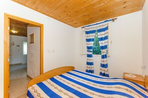 Un dormitorio con techo de madera, una cama con colcha a rayas, una ventana con cortinas azules y blancas, y una mesita de noche con un objeto decorativo.